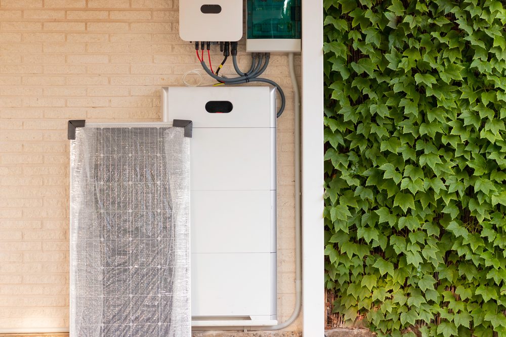 Stacked home battery modules below a hybrid inverter, next to a new solar panel still in its wrapping